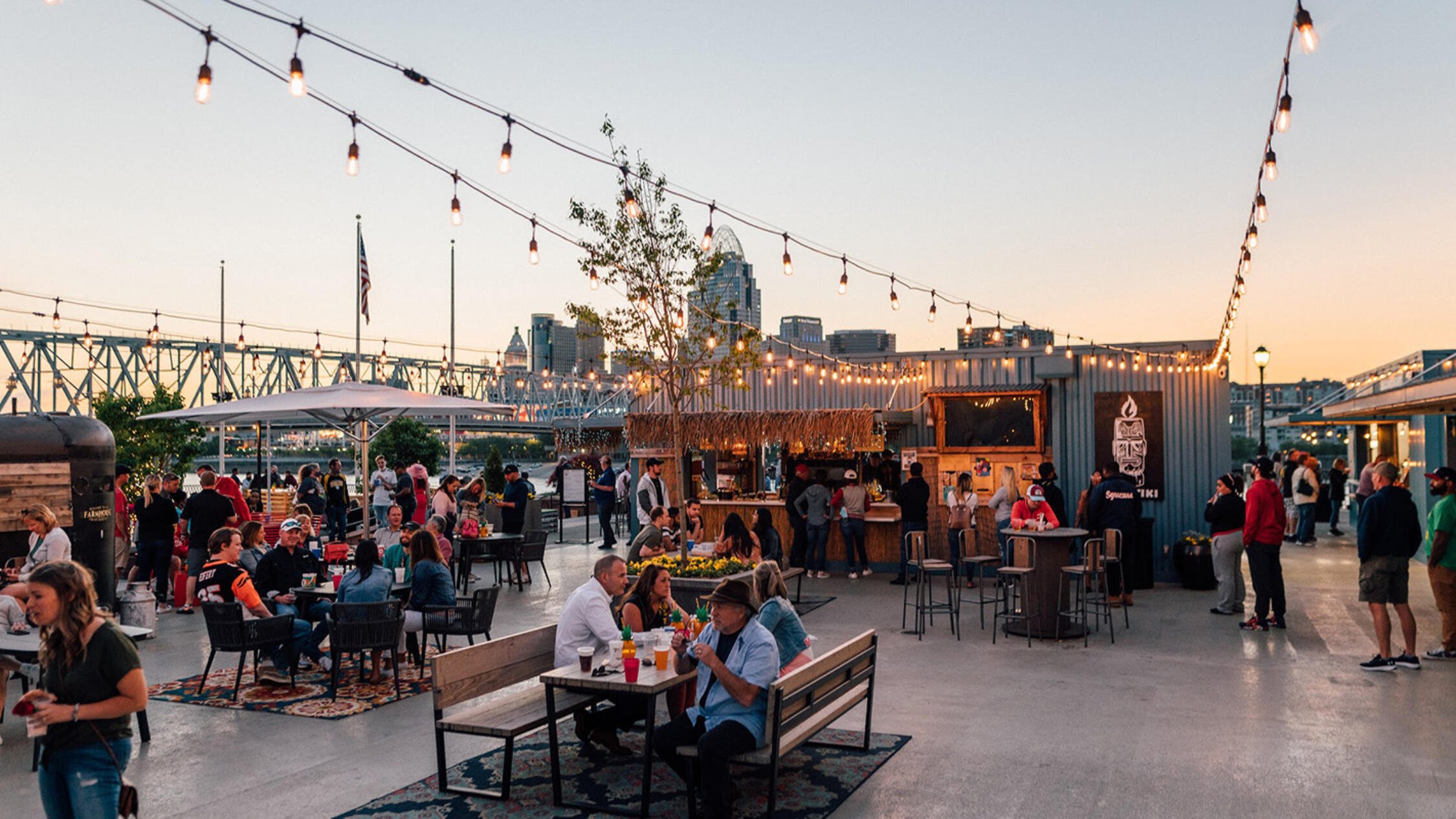 A crowd gathered at an outdoor bar's seating area at Newport on the Levee at dusk with the city skyline in the background