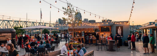 A crowd gathered at an outdoor bar's seating area at Newport on the Levee at dusk with the city skyline in the background